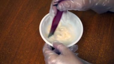 A woman holds a cup with hair dye in her hands and stirs it.