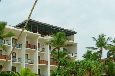 Beyaz bulutlu gökyüzüne karşı otel binası ve palmiye ağaçları.