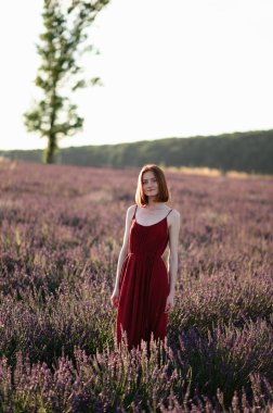 A red-haired young girl without make-up is resting in a lavender field. Summer vacation and travel time.