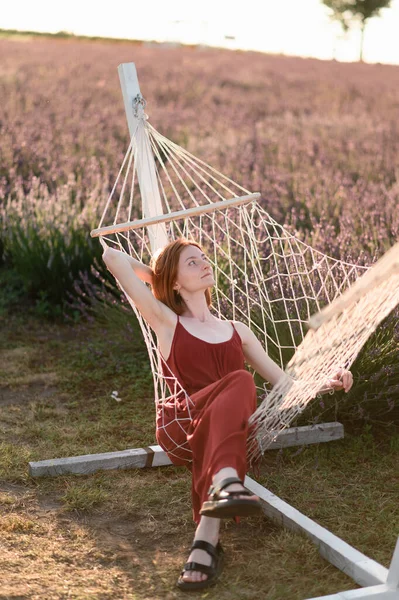 化粧のない赤い髪の少女がラベンダー畑のハンモックに腰を下ろしている 夏休みと旅行時間 — ストック写真