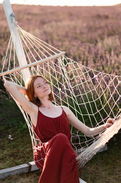化粧のない赤い髪の少女がラベンダー畑のハンモックに腰を下ろしている 夏休みと旅行時間 — ストック写真