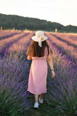 Cheerful young girl in a straw hat and pink dress on the background of a lavender field. Sunset. The girl goes towards the horizon