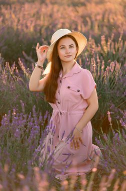 A cheerful young girl in a straw hat and a pink dress sits and rests among lavender bushes. Sunset.