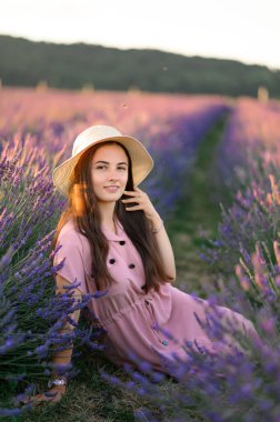 A cheerful young girl in a straw hat and a pink dress sits and rests among lavender bushes. Sunset.