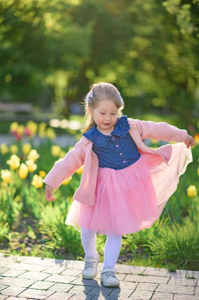 3歳の女の子がドレスとピンクのセーターで公園を歩いています 夏時間だ 赤ん坊はチューリップの背景に彼女のドレスを示しています — ストック写真