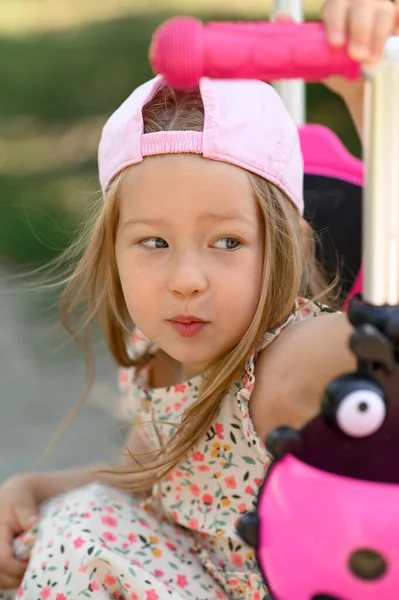 ピンクの帽子の女の子3歳はスクーターに乗っています 夏時間だ 閉じろ ストックフォト
