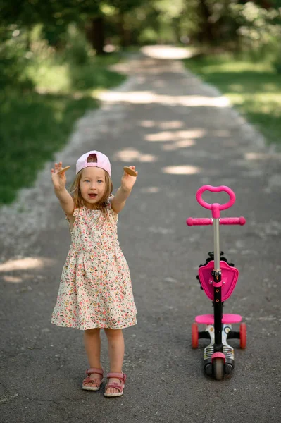 ピンクの帽子の女の子3歳はスクーターに乗っています 夏時間だ 女の子はクッキーを見せます ストックフォト