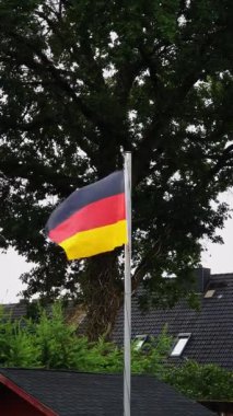 Bir Alman bayrağı bayrak direğinde dalgalanıyor. Arkasında büyük bir ağaç ve yerleşim çatıları olan bulutlu bir günde..