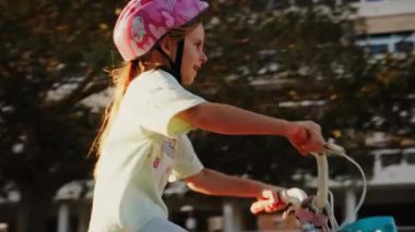 Pembe kask takan genç bir kız heyecandan bisikletine biniyor. Sıcak güneş ışığı hareketlerini vurgular, dinamik ve neşeli bir sahne yaratır..