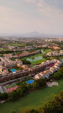 İspanya, Alicante 'de konut binaları, yüzme havuzları ve spor salonlarıyla çevrili canlı bir golf sahasının insansız hava aracıyla ele geçirilmesi. Görüntü, Akdeniz mimarisi, yemyeşil ve şehir planının altın saat boyunca net bir görüntüsünü sunuyor..