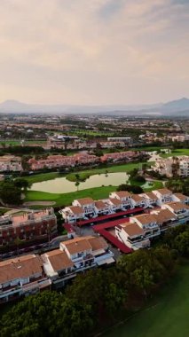 İspanya 'nın Alicante kentinde göletleri ve kırmızı çatılı villaları olan bir golf sahasının yakınındaki Akdeniz tarzı bir mahalleye güzel bir drone perspektifi. Yumuşak bir akşam ışığı bu banliyö sahnesine sıcaklık ve sinematik bir hava katar..