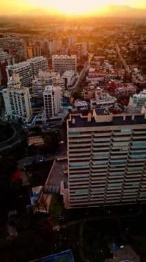 İspanya 'nın Alicante kentinde yüzme havuzu olan Akdeniz tarzı bir konut kompleksinin sinematik hava görüntüsü. İnsansız hava aracı yavaşça yükseliyor, yüksek binaların ve uzak dağların arkasındaki ılık günbatımını ortaya çıkarıyor.