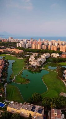 Alicante 'nin sahil şeridini, golf sahalarını ve şehir binalarını gösteren geniş açılı bir fotoğraf. Göletler ve palmiye ağaçları altın saat boyunca Akdeniz atmosferini yansıtarak doğayı kentsel zarafetle harmanlıyor..
