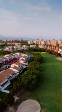 İspanya 'nın Alicante kentindeki yerleşim binalarıyla çevrili manzaralı bir golf sahasının insansız hava aracı çekimi. Görüntü yeşil çayırları, kırmızı tenteli beyaz villaları ve arka planda dağlar olan uzak şehir siluetini yakalıyor.. 