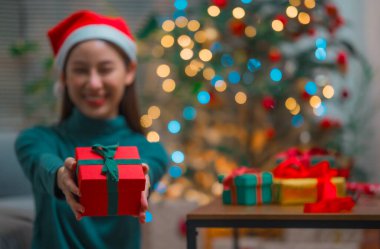 Mutlu gülümseyen Asyalı kadın, Noel Baba şapkalı, yeni yıl hediye kutusunda arka planda Noel ağacı olan.