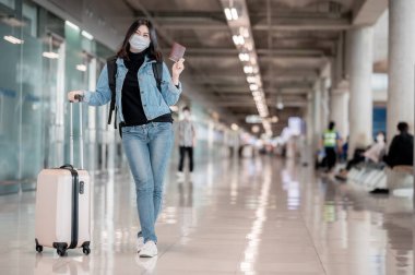 Asyalı gezgin kadın pasaportunu gösteriyor ve uluslararası havaalanında bavullu koruyucu maske takıyor.