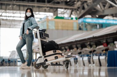 Asyalı gezgin kadın uluslararası havaalanında bebek arabasıyla koruyucu maske takıyor.