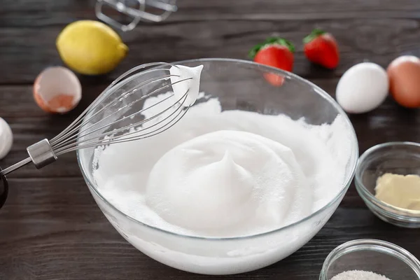 Close-up of a whisk and whipped egg whites to stiff peaks, a step in the preparation of breakfast dough or tender pancakes. White whipped foam of egg whites, bakery preparation. Wooden table