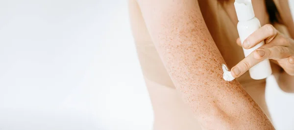 Banner of a woman applying cream on her body, skin protection from sun, skin moisturizing, skincare after shower and bath, treatment. Tanned skin with freckles on a light background, isolated
