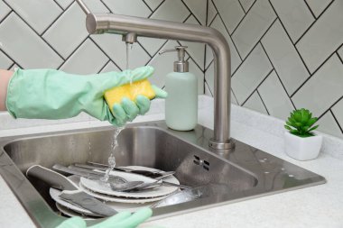 Yeşil eldiven, musluğun altında sünger. Tabaklar ve çatal bıçak takımı lavaboda temizliği bekliyor. Temizlik, ev işleri.