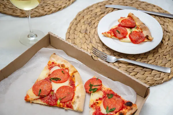 Casual dining setup featuring slices of pepperoni pizza on a plate and in a takeout box, paired with a glass of white wine.