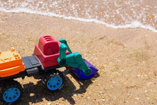 A toy excavator sits on sand, waves in the background. It\'s colorful, playful, family vacation, development crisis, safety rules near sea