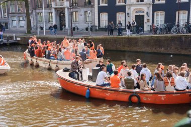 Canlı turuncu renkteki parti katılımcıları bir teknede kutlama yapıyorlar, gülümseyip el sallıyorlar, kanaldaki Kral Bayramı şenlikleri Hollanda, Amsterdam, 27.04.2023