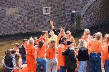 Canlı turuncu renkteki parti katılımcıları bir teknede kutlama yapıyorlar, gülümseyip el sallıyorlar, kanaldaki Kral Bayramı şenlikleri Hollanda, Amsterdam, 27.04.2023