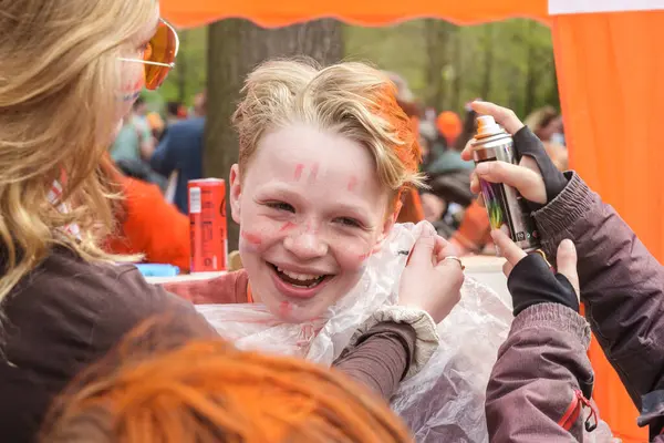 Bir çocuk turuncu saç boyası ve turuncu saç spreyiyle gülüyor. Krallar Günü şenlikleri sırasında Hollanda, Amsterdam, 27.04.2023 'te bir parkta harika vakit geçiriyor.