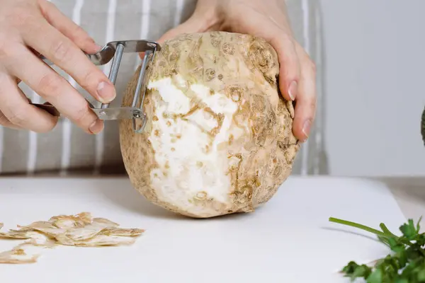 stock image A woman in apron peeling celeriac or celery root with peeler in a kitchen. Celeriac in soups, stews, and healthy recipes rich in antioxidants and minerals