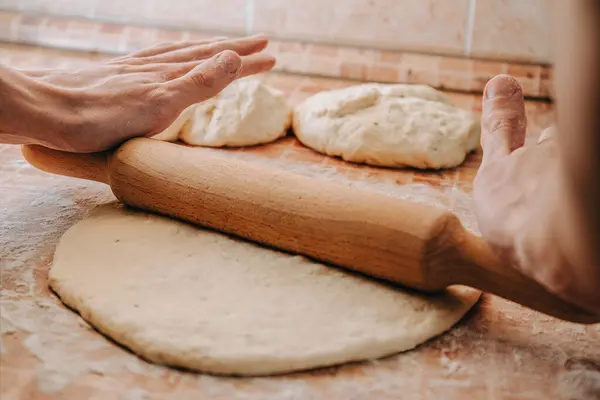 Elleri hamuru tahta bir oklavayla yuvarlıyor. Un kaplı bir yüzeyde arka planda iki hamur topu var..