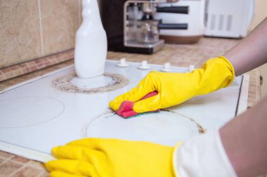 Sarı eldiven takan eller kirli bir mutfak sobasını süngerle, temizlikle ve ev işleriyle temizliyor.