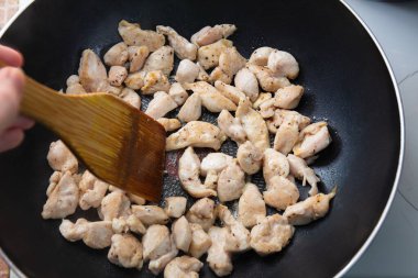 Tavuk parçaları, ahşap bir spatulayla siyah bir tavada pişiriliyor..