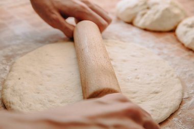 Elleri ahşap bir oklava kullanarak un kaplı bir yüzeyde hamuru düzleştiriyor, pişirmeye hazırlanıyor, ev yapımı ekmek veya pizza yapıyor.