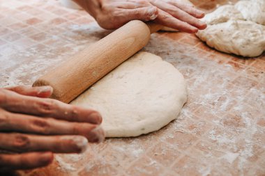 Elleri hamuru ahşap bir oklamayla un kaplı bir yüzeyde yuvarlıyor, hamur pişirme işlemine hazırlanıyor.