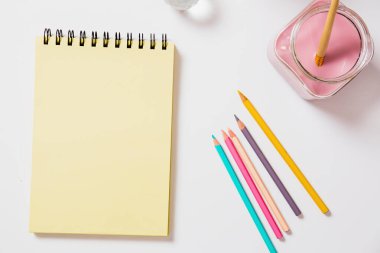 Renkli kalemlerin ve pembe boya kavanozunun yanında boş bir defter var.