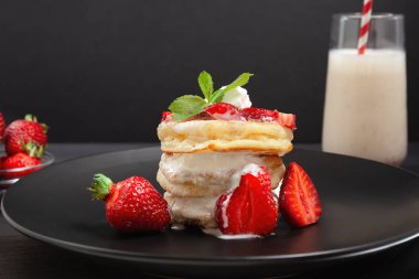Taze çilek ve kremayla kaplı bir yığın pofuduk krep, siyah bir tabakta bir bardak milkshake ile servis edilir..