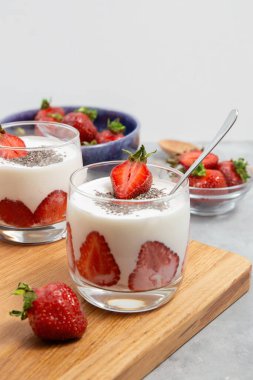 İki bardak çilekli yoğurt üzerine chia tohumu serpiştirilmiş. Tahta bir tahtanın üzerinde taze çilek ile servis ediliyor..