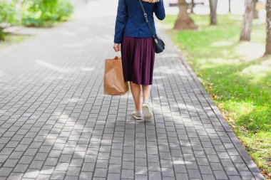 Kahverengi bir alışveriş çantası taşıyan, parktaki kaldırımlı bir yolda yürüyen bir kadının arka görüntüsü.