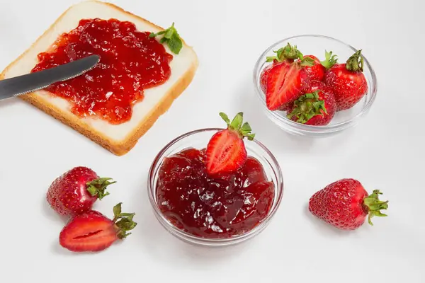 stock image Slice of bread toast with strawberry jam spread, fresh strawberries, and bowls of jam on a white background