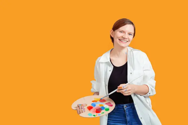 stock image Smiling woman in a white shirt and jeans holding a paint palette and brush, standing against an solid orange background