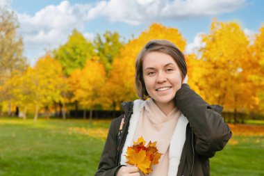 Sıcak bir gülümsemesi olan genç bir kadın, sonbahar yapraklarını tutar, arka planda parlak sarı ağaçlar olan bir parkta durur.