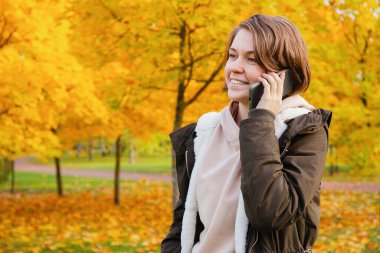 Sıcak ceketli gülümseyen kadın, canlı bir sonbahar yeşilliği olan bir parkta dururken telefonda konuşmaktan zevk alıyor.