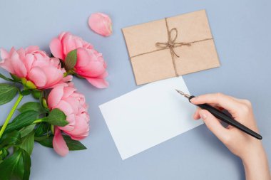 Elinde bir kalem var, boş bir karta yazmaya hazır pembe çiçekler, bir zarf ve pastel mavi arka planda yapraklar.