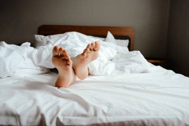 Sıcak bir yatak odasında, beyaz yatak çarşaflarının altından sarkan, rahat ve tembel bir sabah ya da uyanan bir insan.