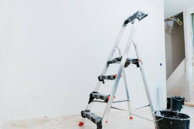 İnşaat merdiveni, kısmen yenilenmiş boş beyaz duvarlı bir odada boya kovalarının yanında duruyor.