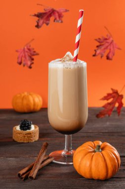 Pumpkin spice latte in a tall glass with foam, cinnamon, pumpkins, and falling leaves in a cozy fall setting on a wooden table clipart