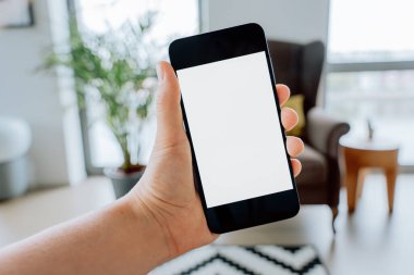 Bir kişi, içi bulanık, modern oturma odasının içinde boş beyaz bir ekranla dikey olarak akıllı bir telefonu tutar.