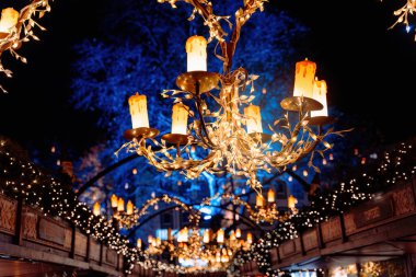 Chandeliers with warm candle lights hang above a festive Christmas market, creating a magical glow under the night sky clipart