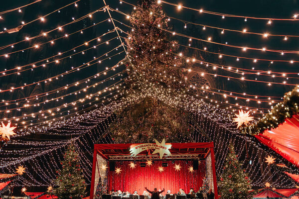 Germany, Cologne, 12.10.2022 - Christmas market concert stage under festive lights, featuring large decorated tree and red market stalls